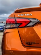 The rear section of an orange car with the model name VERSA prominently displayed. The taillight features a modern, angular design. There's a logo or website URL, 'EdMartin.com', printed near the bottom. The background reveals a cloudy sky and a blue car parked nearby.