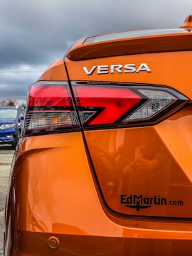 The rear section of an orange car with the model name VERSA prominently displayed. The taillight features a modern, angular design. There's a logo or website URL, 'EdMartin.com', printed near the bottom. The background reveals a cloudy sky and a blue car parked nearby.