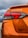 The rear section of an orange car with the model name VERSA prominently displayed. The taillight features a modern, angular design. There's a logo or website URL, 'EdMartin.com', printed near the bottom. The background reveals a cloudy sky and a blue car parked nearby.
