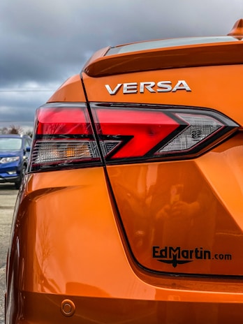 The rear section of an orange car with the model name VERSA prominently displayed. The taillight features a modern, angular design. There's a logo or website URL, 'EdMartin.com', printed near the bottom. The background reveals a cloudy sky and a blue car parked nearby.