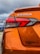 The rear section of an orange car with the model name VERSA prominently displayed. The taillight features a modern, angular design. There's a logo or website URL, 'EdMartin.com', printed near the bottom. The background reveals a cloudy sky and a blue car parked nearby.