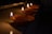 A row of lit oil lamps, known as diyas, is placed on a flat surface in a dimly lit environment. The flames flicker gently, casting a warm glow and creating a serene and peaceful atmosphere.