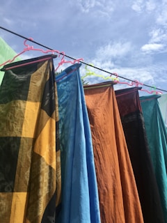 Several colorful fabrics hang on a clothesline, each held by a different colored hanger. The fabrics display various patterns and textures, including a black and yellow checkered design, solid blue, solid orange, and others with intricate designs. The scene is outdoors, under a bright, partly cloudy sky.