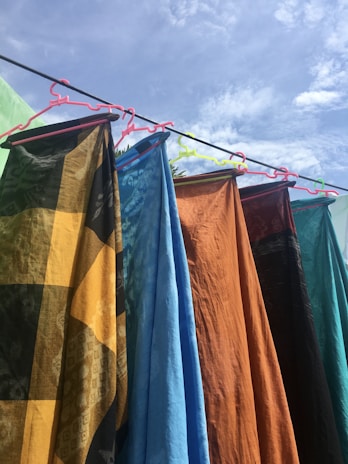 Several colorful fabrics hang on a clothesline, each held by a different colored hanger. The fabrics display various patterns and textures, including a black and yellow checkered design, solid blue, solid orange, and others with intricate designs. The scene is outdoors, under a bright, partly cloudy sky.