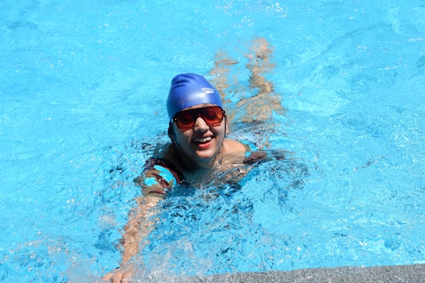 A person is swimming in a pool, wearing a blue swim cap and red goggles. The individual appears to be smiling and is partially submerged in bright blue water, creating splashes around them.