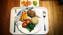 A well-arranged meal is on a white tray on a wooden table, with a plate featuring lamb chops, a serving of brown rice, grilled tomatoes, saut&eacute;ed greens, and sliced yellow vegetables. On a small side plate are shrimp with onions and a portion of guacamole. Cutlery is placed neatly beside the plate.