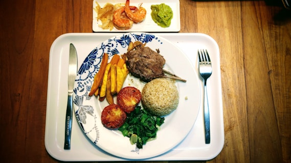 A well-arranged meal is on a white tray on a wooden table, with a plate featuring lamb chops, a serving of brown rice, grilled tomatoes, sautéed greens, and sliced yellow vegetables. On a small side plate are shrimp with onions and a portion of guacamole. Cutlery is placed neatly beside the plate.