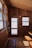 A cozy wooden interior with a ceiling, walls, and floor crafted from wood. There is a wooden door with a window, and a separate glass window to the side offering a view outside. A bench and small table are positioned in the corner and a fire extinguisher is mounted on the wall beside the door.