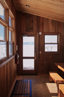 A cozy wooden interior with a ceiling, walls, and floor crafted from wood. There is a wooden door with a window, and a separate glass window to the side offering a view outside. A bench and small table are positioned in the corner and a fire extinguisher is mounted on the wall beside the door.