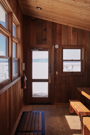 A cozy wooden interior with a ceiling, walls, and floor crafted from wood. There is a wooden door with a window, and a separate glass window to the side offering a view outside. A bench and small table are positioned in the corner and a fire extinguisher is mounted on the wall beside the door.