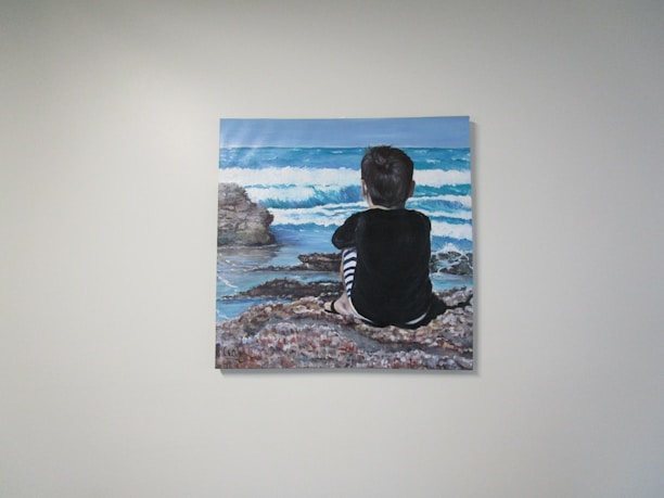 A painting on a wall depicts a child sitting on a rocky shoreline, gazing at the ocean waves. The child is wearing a black top with striped pants, and the scene conveys a sense of contemplation and tranquility.