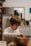 A child wearing a striped shirt intently mixes something in a large metal bowl. The scene appears to be set in a cozy, homey kitchen with a wooden table and some blurred background elements such as plants and framed pictures.