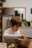 A child wearing a striped shirt intently mixes something in a large metal bowl. The scene appears to be set in a cozy, homey kitchen with a wooden table and some blurred background elements such as plants and framed pictures.