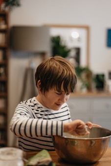 A child wearing a striped shirt intently mixes something in a large metal bowl. The scene appears to be set in a cozy, homey kitchen with a wooden table and some blurred background elements such as plants and framed pictures.