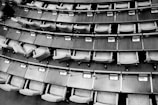Rows of empty seats and wooden desks arranged in a curved layout, typically found in a legislative assembly or lecture hall setting. Each desk has a small electronic device or voting panel.
