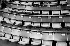 Rows of empty seats and wooden desks arranged in a curved layout, typically found in a legislative assembly or lecture hall setting. Each desk has a small electronic device or voting panel.
