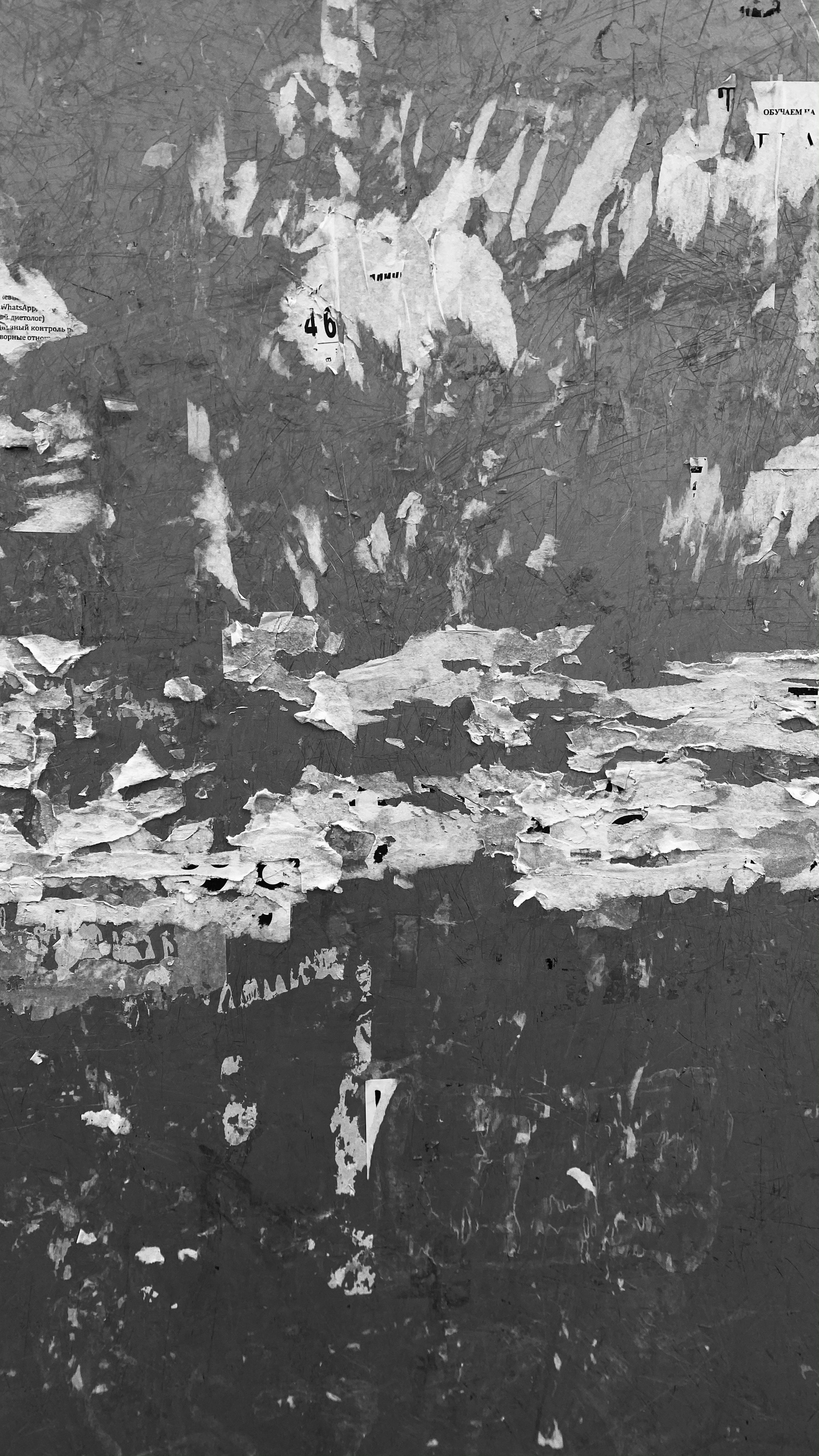 A textured wall surface with layers of peeling paint and small pieces of torn paper. The surface is rough, with patches of different shades and no discernible pattern. Random letters and numbers are scattered across the image.