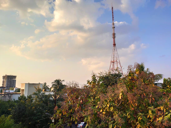 A tall communication tower stands prominently against a sky filled with clouds, positioned amidst trees and foliage. In the background, a partially constructed high-rise building can be seen, along with a few smaller structures. The image captures a blend of urban elements with natural greenery.