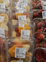 Plastic containers hold mixed fruit slices such as pineapples, oranges, and grapes, next to packages of fresh strawberries. Price labels indicate the cost per kilogram and emphasize immediate consumption.