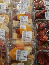 Plastic containers hold mixed fruit slices such as pineapples, oranges, and grapes, next to packages of fresh strawberries. Price labels indicate the cost per kilogram and emphasize immediate consumption.