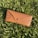A brown textured eyeglass case with a Ray-Ban logo is lying on freshly mowed green grass, basking in sunlight.