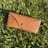 A brown textured eyeglass case with a Ray-Ban logo is lying on freshly mowed green grass, basking in sunlight.