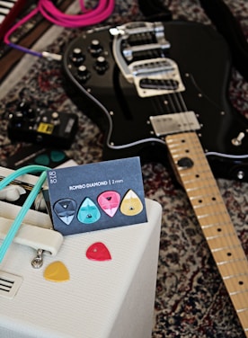 A black electric guitar is laying on a patterned carpet alongside a collection of colorful guitar picks displayed on a card labeled 'ROMBO DIAMOND | 2 mm'. A white amplifier with retro design details and a turquoise cable is positioned nearby, while an effects pedal can be seen on the carpet.