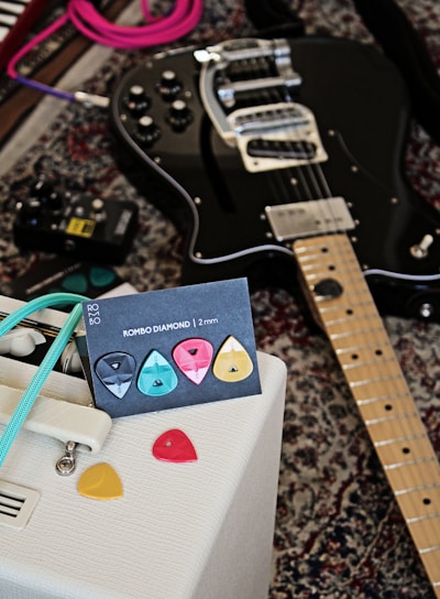 A black electric guitar is laying on a patterned carpet alongside a collection of colorful guitar picks displayed on a card labeled 'ROMBO DIAMOND | 2 mm'. A white amplifier with retro design details and a turquoise cable is positioned nearby, while an effects pedal can be seen on the carpet.