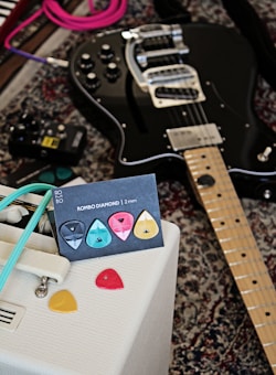 A black electric guitar is laying on a patterned carpet alongside a collection of colorful guitar picks displayed on a card labeled 'ROMBO DIAMOND | 2 mm'. A white amplifier with retro design details and a turquoise cable is positioned nearby, while an effects pedal can be seen on the carpet.