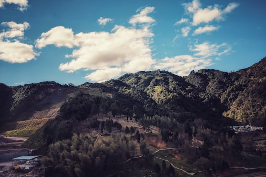 A picturesque landscape featuring lush green mountains under a bright blue sky with scattered white clouds. There are dense forests covering the mountains and patches of grass and pathways visible. The sunlight casts shadows creating a mix of light and dark areas on the terrain.
