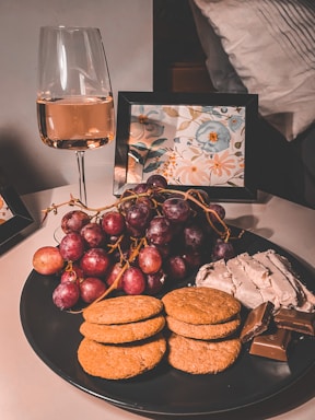 A cozy and elegant arrangement featuring a glass of rose wine, a plate with a selection of red grapes, crackers, cheese, and chocolate. The background includes a partly visible floral artwork in a frame positioned on a table next to a pillow.