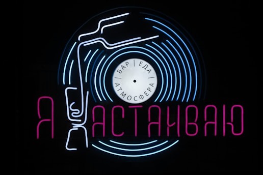 A neon sign featuring a stylized vinyl record with a tonearm and text in Cyrillic script. The sign uses vibrant blue and pink colors against a black background, creating a visually striking and retro aesthetic. The words 'бар', 'еда', and 'атмосфера' are written on the record label, hinting at a bar, food, and atmosphere theme.