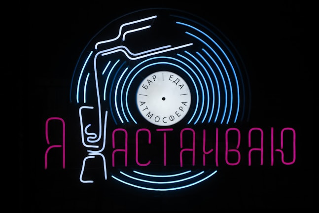 A neon sign featuring a stylized vinyl record with a tonearm and text in Cyrillic script. The sign uses vibrant blue and pink colors against a black background, creating a visually striking and retro aesthetic. The words 'бар', 'еда', and 'атмосфера' are written on the record label, hinting at a bar, food, and atmosphere theme.