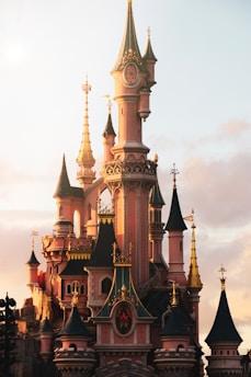 A fairy tale castle with multiple turrets and spires, adorned with gold details and a mix of pink and blue tones. The design features intricate architectural elements such as pointed towers and decorative trim, capturing the glow of the setting sun.