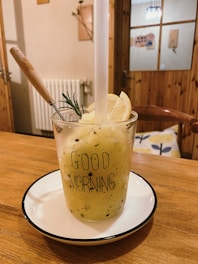 A glass filled with a yellow smoothie, garnished with a slice of lemon and a sprig of rosemary, sits on a white plate with a black rim. The glass has 'Good Morning' printed on it. A wooden spoon is placed inside the drink. The setting appears to be a cozy, wooden-furnished room with visible wooden chairs, a door, and some decorations on the walls.