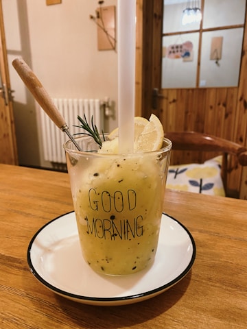 A glass filled with a yellow smoothie, garnished with a slice of lemon and a sprig of rosemary, sits on a white plate with a black rim. The glass has 'Good Morning' printed on it. A wooden spoon is placed inside the drink. The setting appears to be a cozy, wooden-furnished room with visible wooden chairs, a door, and some decorations on the walls.