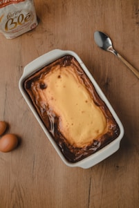 A freshly baked cheesecake is placed in a white rectangular baking dish on a wooden surface. Next to it, there are two brown eggs, a spoon, and a bag of flour. The cheesecake has a golden-brown crust around the edges and a creamy yellow center.