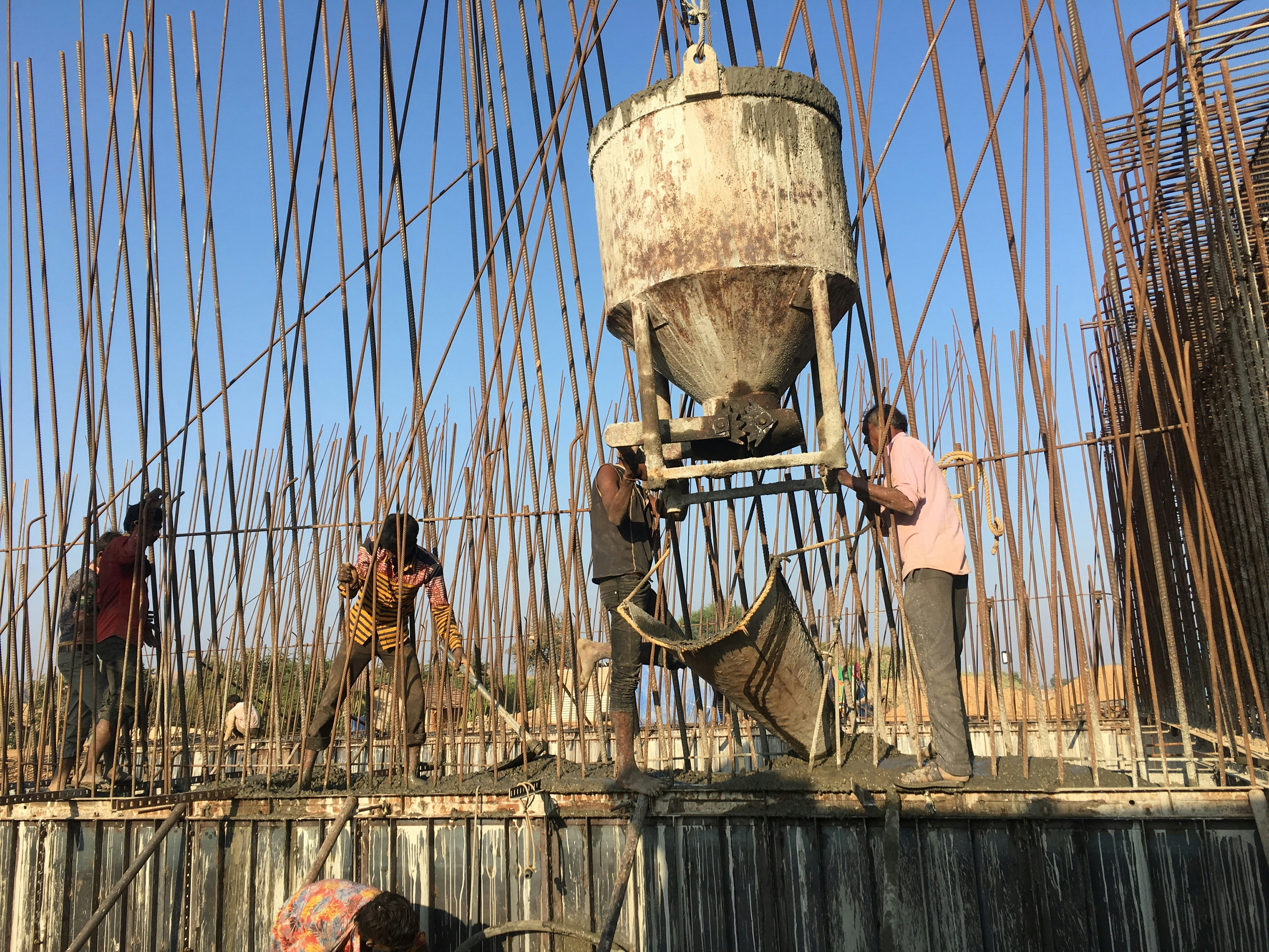 Several workers are engaged in a construction project, surrounded by numerous rebar rods. They are using equipment to pour concrete, with one worker guiding the chute and others spreading the mixture on the surface.