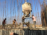 Several workers are engaged in a construction project, surrounded by numerous rebar rods. They are using equipment to pour concrete, with one worker guiding the chute and others spreading the mixture on the surface.