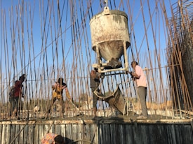 Several workers are engaged in a construction project, surrounded by numerous rebar rods. They are using equipment to pour concrete, with one worker guiding the chute and others spreading the mixture on the surface.