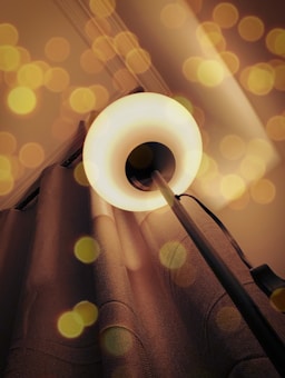 A floor lamp with a circular light source casts a warm glow, surrounded by bokeh effects. The lamp stands in front of textured curtains, creating a cozy ambiance.