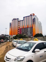 A large, colorful multi-story building features a pattern of red, orange, black, and white. In front of the building, a row of white taxis waits along the side of a road, with a grey concrete wall partially visible on one side and trees in the background.