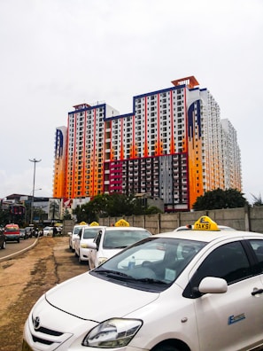 A large, colorful multi-story building features a pattern of red, orange, black, and white. In front of the building, a row of white taxis waits along the side of a road, with a grey concrete wall partially visible on one side and trees in the background.