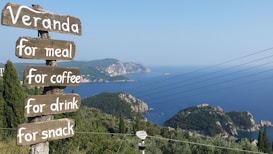 A scenic coastal view with a series of wooden signboards. The signs read 'Veranda', 'for meal', 'for coffee', 'for drink', and 'for snack'. The background features lush green hills leading to a vast blue sea, and the sky is clear with a hint of haze. Power lines stretch across the scene, adding an element of modernity to the natural setting.