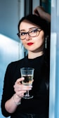 A woman with long dark hair and glasses is holding a glass of white wine. She is wearing a black top and red lipstick, standing with one arm resting above her head against a doorway. The setting is indoors with natural light coming from the side.