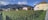 A picturesque vineyard stretches across the foreground, lush with rows of green grapevines. In the background, charming houses with traditional architecture can be seen. Beyond the houses, a range of majestic mountains rises, partially covered in snow, under a sky spotted with clouds.