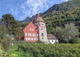 A picturesque red and stone building with a tall, pointed roof is set against a backdrop of lush green trees and a mountain range. The architecture features white-framed windows and decorative brickwork. The foreground includes neatly arranged grapevines and a grassy area, suggesting a vineyard setting. A bright blue sky with some clouds completes the scenic view.