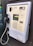 A wall-mounted public payphone with a handset and metal cord. The panel includes a coin slot, a keypad with buttons marked for various services like last number redial and volume control, and a coin return slot at the bottom.