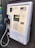 A wall-mounted public payphone with a handset and metal cord. The panel includes a coin slot, a keypad with buttons marked for various services like last number redial and volume control, and a coin return slot at the bottom.