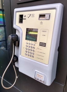 A wall-mounted public payphone with a handset and metal cord. The panel includes a coin slot, a keypad with buttons marked for various services like last number redial and volume control, and a coin return slot at the bottom.